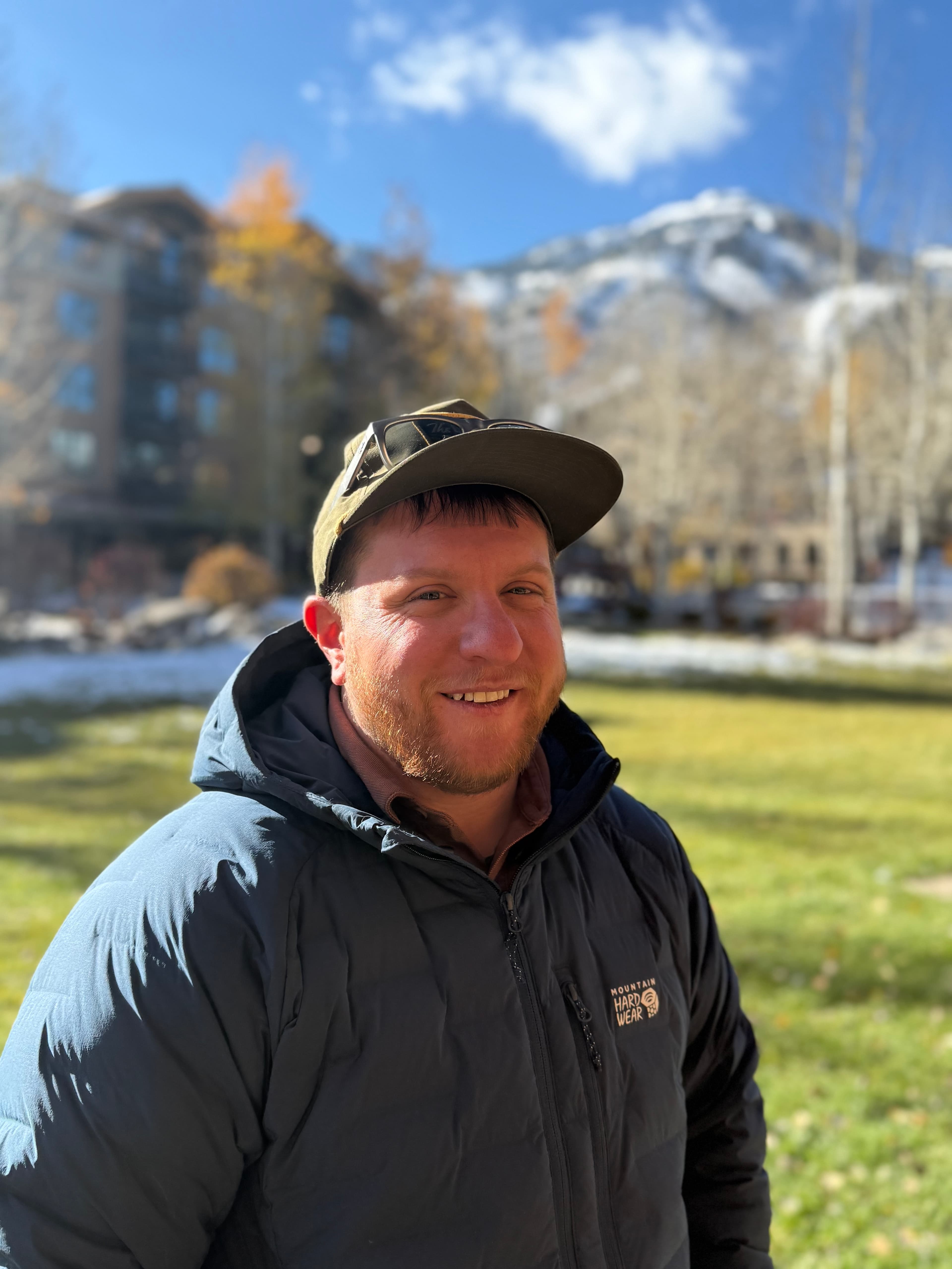 Smiling man in a blue puffer jacket and cap with snowy mountains in the background.